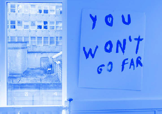 A blue and white toned photograph which, on the right, shows a white board with black scrawled writing which reads ‘YOU WON’T GO FAR’. It hangs at a jaunty angle against a white wall. On the left of the image, a window looks out onto a scene of urban dereliction, featuring waste ground and a building in which windows are smashed.